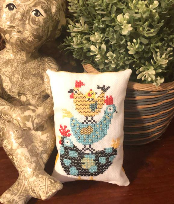 May include: A small, white decorative pillow with a cross-stitched design featuring three chickens in shades of blue, yellow, and red. The chickens are nested in a black and teal basket. The pillow is rectangular and has a soft, plush appearance.